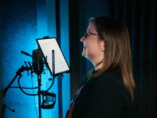 Eine Frau mit Brille lacht und blickt auf ein Tablet, das vor einem Mikrofon steht.
