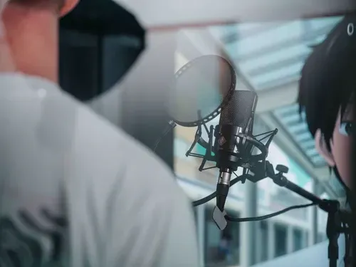 Eine Person in einem Studio sitzt vor einem Mikrofon mit Poppschutz, im Hintergrund ist ein animierter Charakter zu sehen.