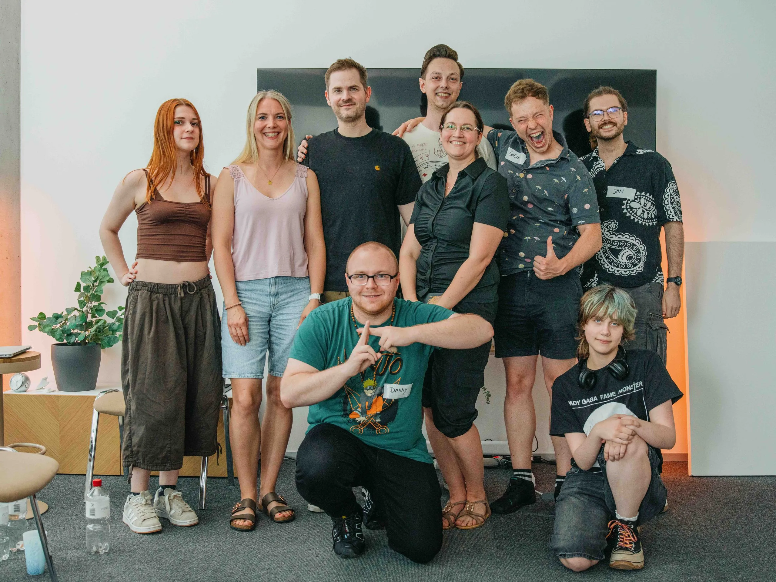 Neun junge Menschen stehen und sitzen für ein Gruppenfoto vor einer Wand.