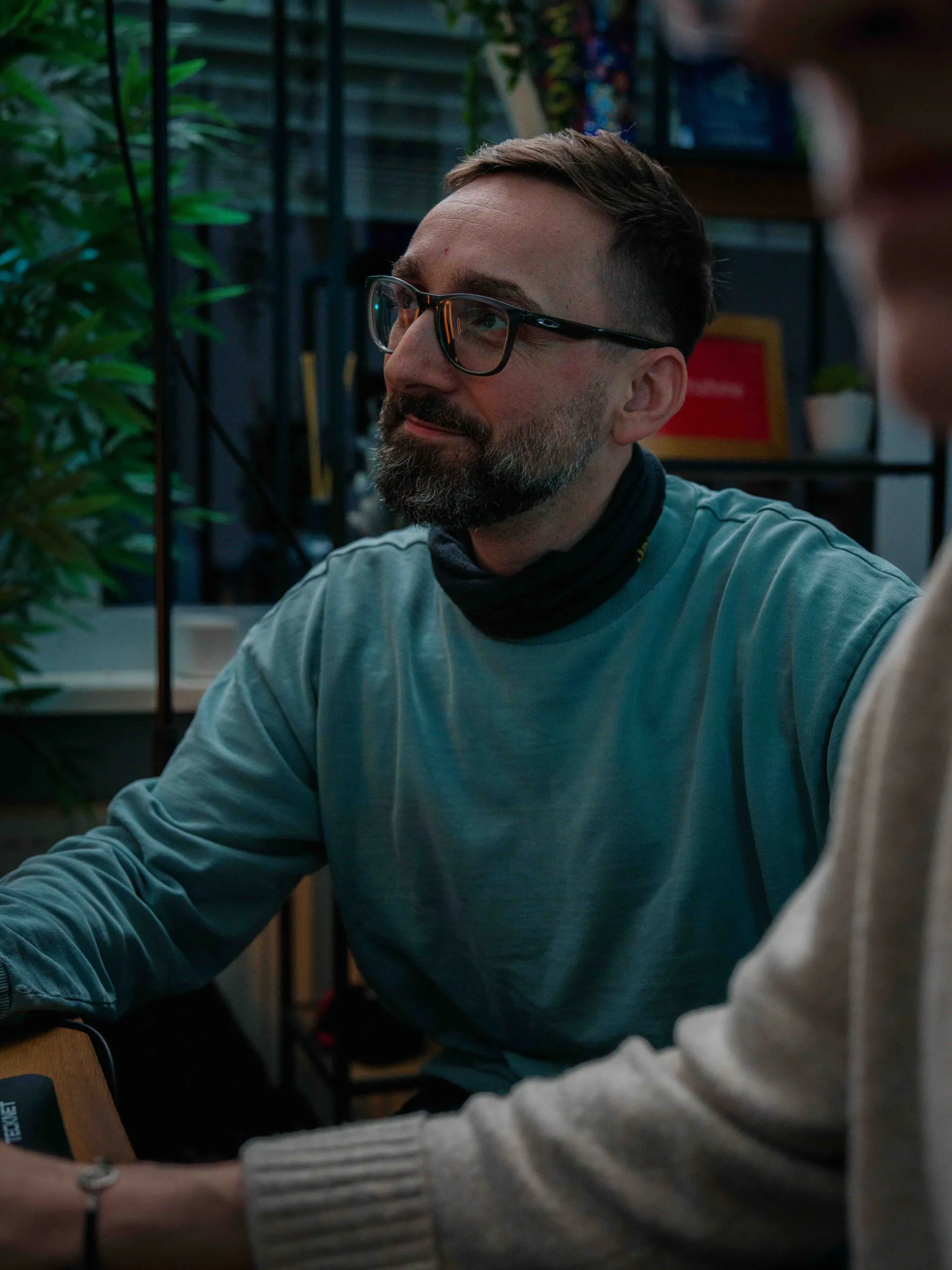 Ein Mann mit Brille und Bart schaut nach links, während er mit einer Person spricht, deren Oberkörper nur unscharf zu sehen ist.