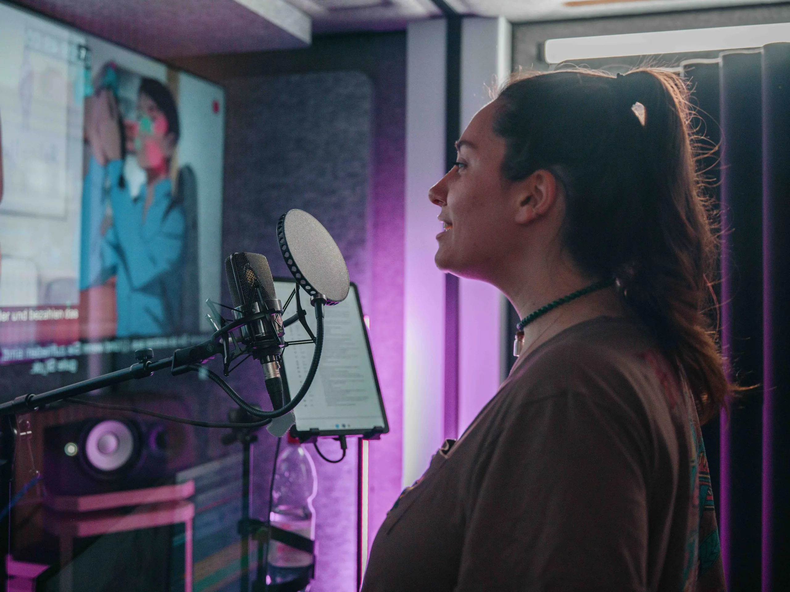 Eine Frau spricht in ein Mikrofon in einem Tonstudio, während auf einem Bildschirm im Hintergrund eine Person zu sehen ist.