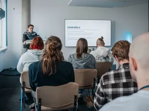Ein Referent steht vor einer Gruppe von Zuhörern und präsentiert eine Folie mit dem Titel "ENSEMBLE".