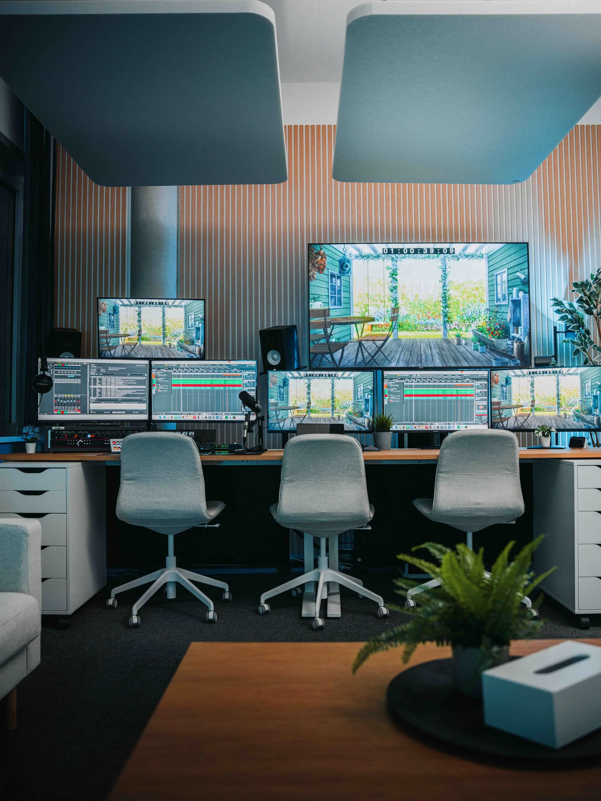 Eine Reihe von Computermonitoren, die für die Postproduktion eingerichtet sind, mit drei grauen Bürostühlen davor.