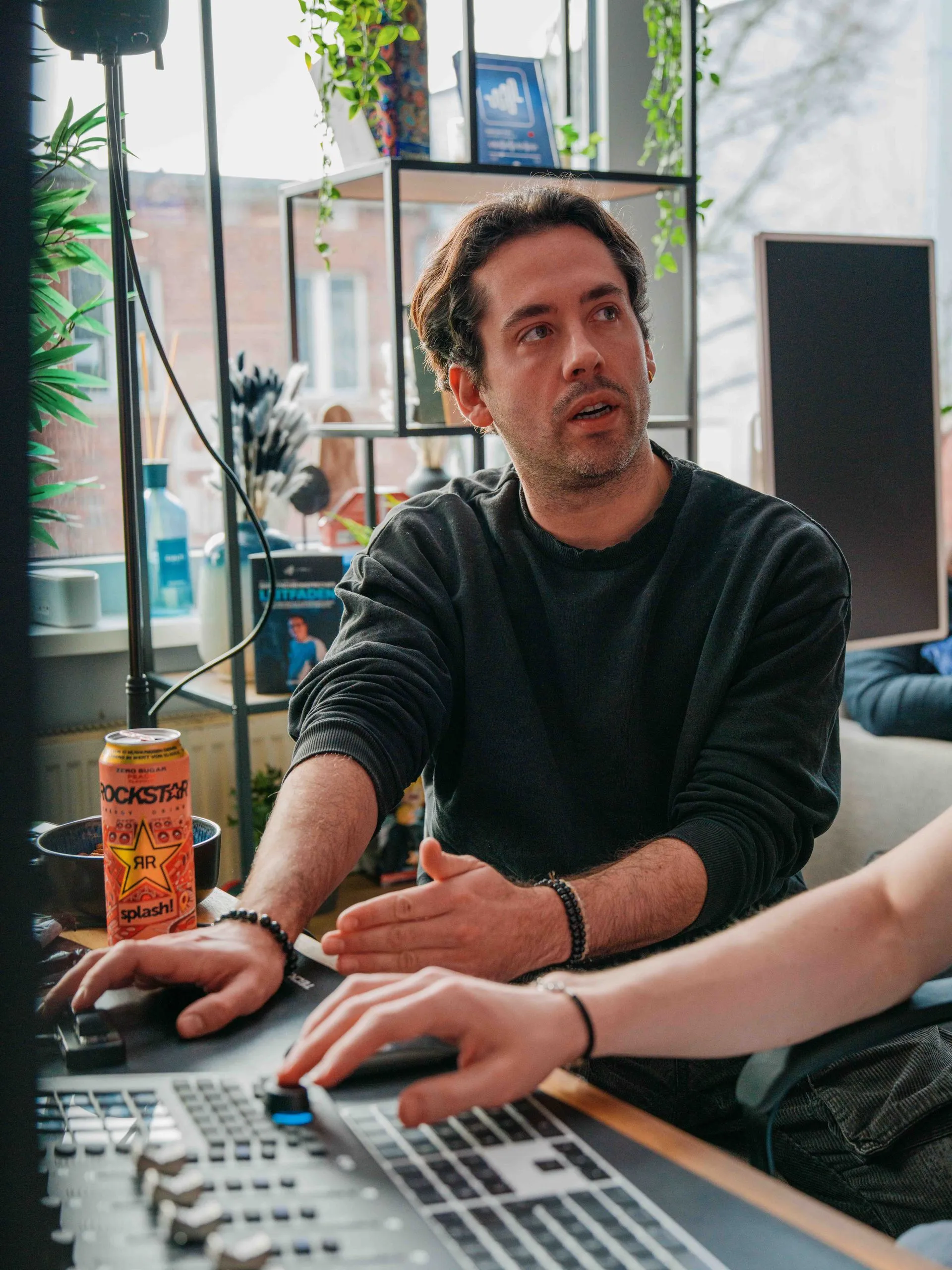 Ein Mann in einem schwarzen Pullover sitzt an einem Mischpult und gestikuliert, während neben ihm eine Dose Rockstar-Energydrink steht.