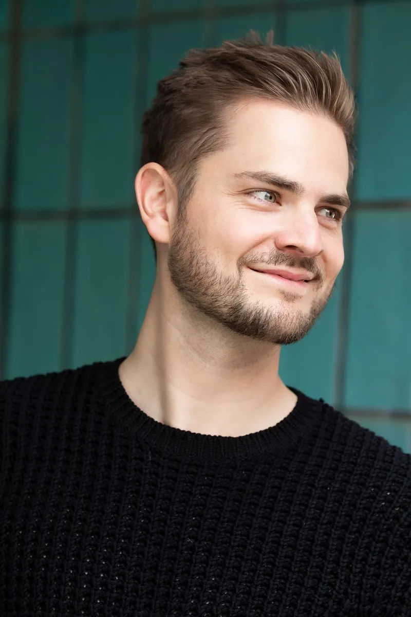 Ein junger Mann mit Bart und braunen Haaren lächelt und schaut nach rechts, er trägt einen schwarzen Pullover vor einem türkisfarbenen Hintergrund mit vertikalen Linien.