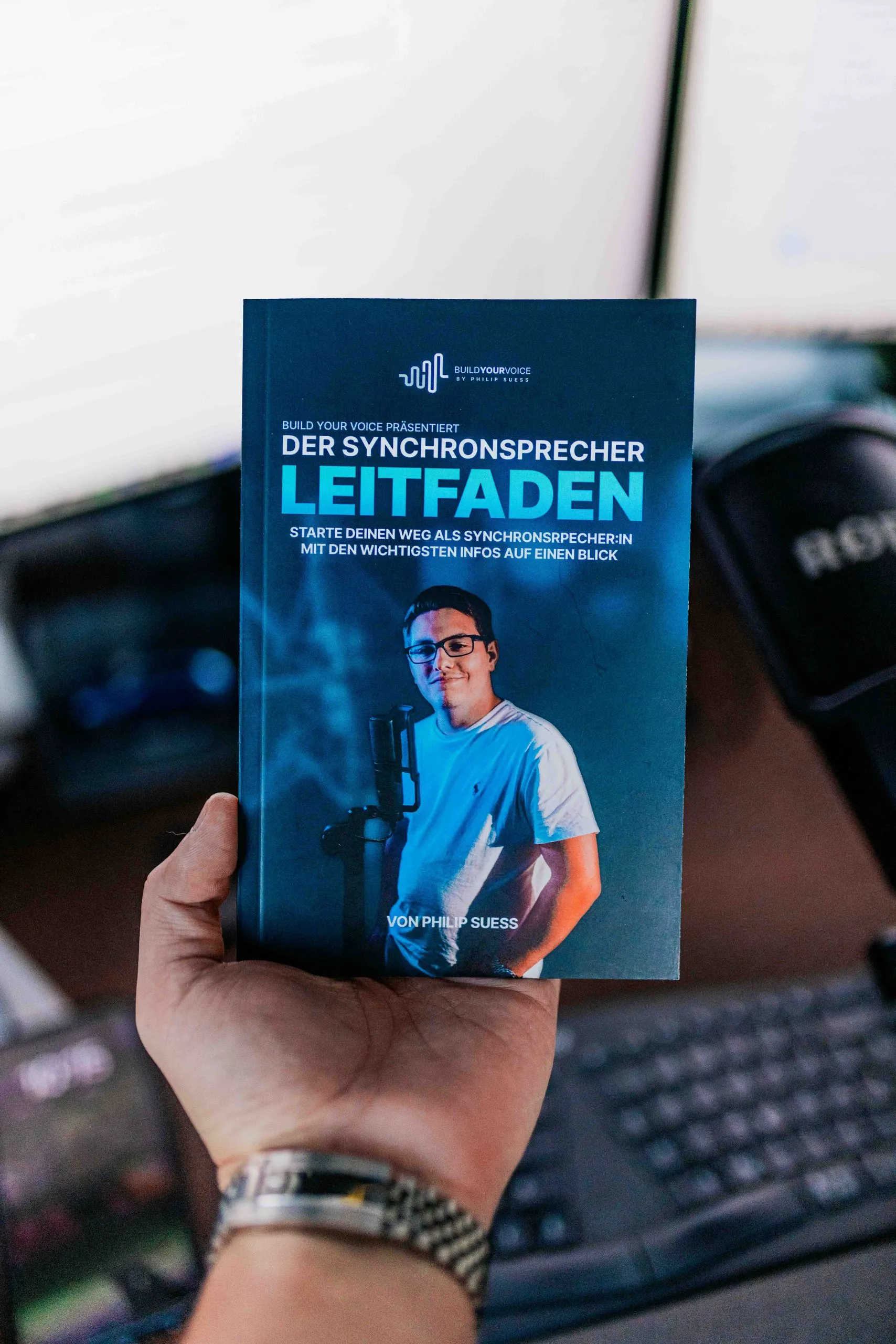 Ein Buch mit dem Titel „Der Synchronsprecher Leitfaden“ von Philip Suess liegt in der Hand, daneben ist ein Mikrofon zu sehen.