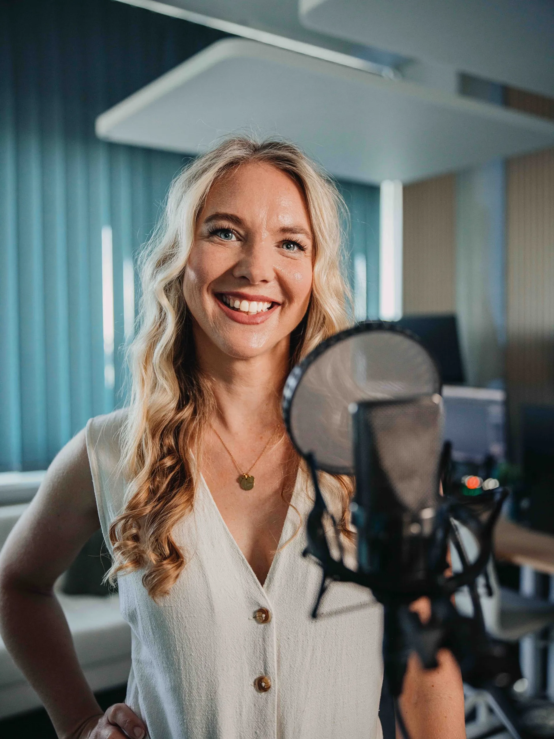 Eine Frau mit blonden Haaren und einem breiten Lächeln steht in einem Aufnahmestudio vor einem Mikrofon.