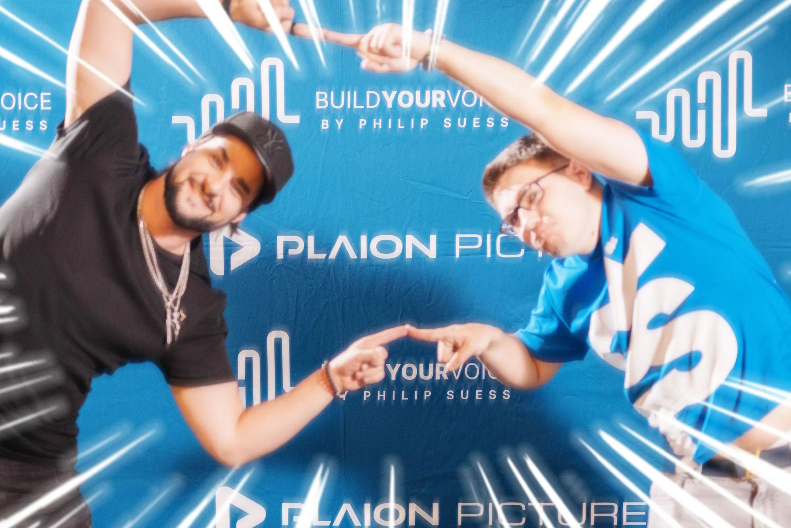 Zwei Männer mit gestreckten Armen und Fingern zueinander bilden einen Rahmen vor einem blauen Hintergrund mit dem Schriftzug "BUILD YOUR VOICE BY PHILIP SUESS" und "PLAION PICTURE".